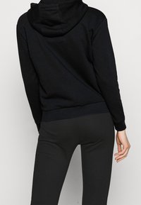 Sudadera negra con un bolsillo canguro y capucha ajustable con cordón. El material parece suave y texturizado, con una cinturilla y puños ajustados.