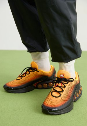 Persona, kas valkā spilgti oranžas un melnas Nike Air Max kedas ar baltām zeķēm un melniem biksēm uz zaļa grīdas.