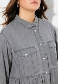 Camicia grigia botton-up realizzata in morbido denim, con due tasche sul petto, bottoni perle, colletto e orlo arricciato per un fit comodo.
