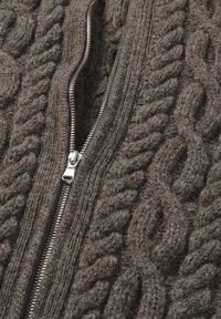 Bruine gebreide trui met een gestructureerd kabelpatroon en een zilveren ritsluiting in het midden. De stof lijkt dik en comfortabel te zijn.
