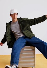 Veste en polaire verte doublée sur une chemise grise, jeans larges bleus et baskets grises, assis sur un tabouret en métal rayé contre un fond blanc.