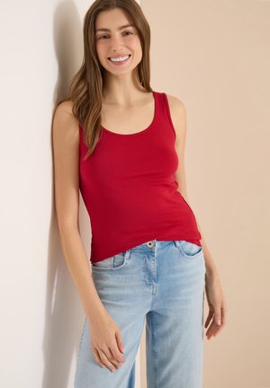 Femme aux longs cheveux bruns sourit, debout contre un mur, portant un haut rouge sans manches et un jean bleu clair.