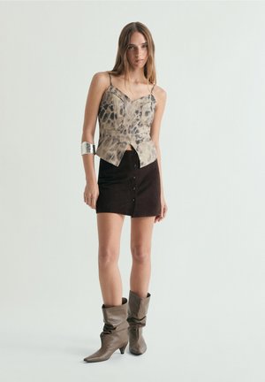 Mujer de pie con una blusa beige con estampado animal, mini falda negra con botones, brazalete plateado y botines taupe holgados.