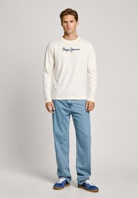 Langärmliges cremefarbenes Hemd mit dem Logo "Pepe Jeans London", kombiniert mit hellblauen Jeans und blauen Sneakern mit weißen Akzenten.