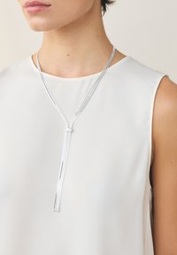 Silberne Metallschmuckkette mit mehreren Strängen, einem zentralen Knoten und fließenden Enden, getragen über einem cremefarbenen, ärmellosen Oberteil.