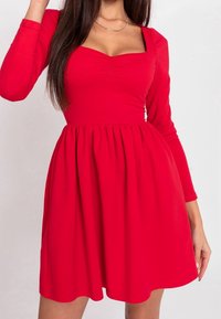 Czerwona sukienka z długim rękawem i kwadratowym dekoltem, z marszczoną górą i rozszerzanym dołem. Gładki materiał o jednolitej teksturze. Brak widocznych wzorów lub elementów metalowych.