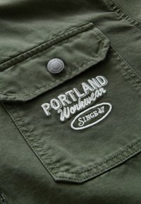 Grüner Stoff mit einer Tasche, die weißen gestickten Text "PORTLAND Workwear" und "Since 47" zeigt, akzentuiert durch einen silbernen Druckknopf.
