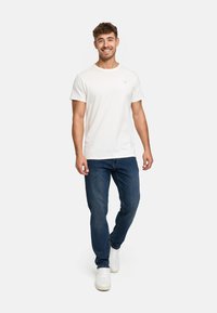 Weißes Baumwoll-T-Shirt mit kurzen Ärmeln, kombiniert mit blauen Denim-Jeans. Das Modell geht und trägt weiße Sneaker. Einfaches Design, lässige Passform.