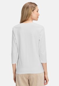 Weißes Langarm-T-Shirt mit Rundhalsausschnitt und drei Viertelärmeln, über beigefarbenen Cordhosen getragen. Glatte Textur, lässige Passform.
