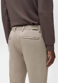 Beige tapered Hosen mit einer glatten Textur, ausgestattet mit Gesäßtaschen und einem elastischen Bund, kombiniert mit einem dunkelbraunen Langarmoberteil.
