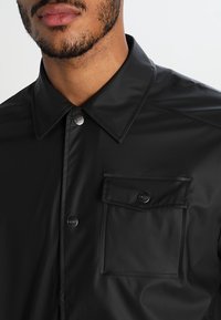 Svart, lätt skjorta med glansig yta, spetsig krage, ficka fram och tryckknappar. Minimalistisk design utan mönster.