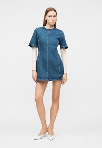 Νεαρή γυναίκα στέκεται, φορώντας μίνι φόρεμα από τζιν με κοντά μανίκια πάνω από άσπρο τοπ με μακριά μανίκια, και τακούνια με λευκή μυτερή μύτη.