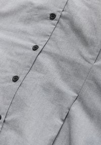 Nærbillede af en lysegrå stofskjorte med sorte knapper og synlig syning langs knappepladsen.