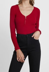 Punainen ribbineulottu bodysuit, jossa pitkät hihat, syvä V-kaula-aukko ja metallivetoketju, yhdistetty korkeavyötäröisiin mustiin farkkuihin.