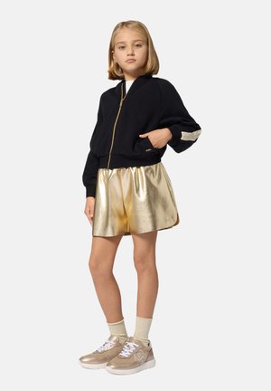 Veste bomber noire zippée avec des accents dorés, jupe métallisée dorée, baskets beiges avec lacets blancs, et chaussettes dorées. Textures lisses et tissu brillant.