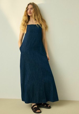 Ung kvinde med langt blondt hår iført en ærmeløs mørkeblå denim maxikjole og sorte flettede sandaler, stående mod en ensfarvet baggrund.