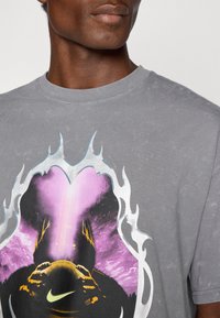 Grå bomulds T-shirt med et farverigt grafisk design, der inkluderer et cirkulært billede, flammer og en central figur, der holder en bold.
