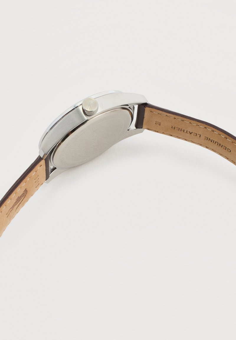 Polshorloge met zilverkleurige kast, leren band met label "Echt leer" en zichtbare kroon, afgebeeld op een witte achtergrond, met de achterkant van het horloge zichtbaar.