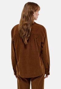 Chemise en velours côtelé marron à manches longues, avec des poignets boutonnés et une coupe décontractée. Sa texture douce et le détail du pli dans le dos rehaussent son design.