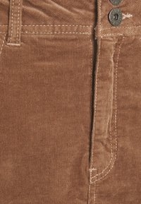 I pantaloni in corduroy marroni presentano costine verticali, una chiusura con bottone e cuciture bianche a contrasto lungo le cuciture e le tasche.