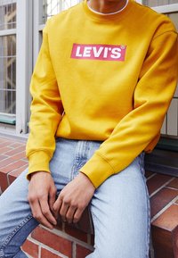 Gelbes Sweatshirt mit einem roten und weißen "LEVI'S"-Logo, Rundhalsausschnitt, gerippte Bündchen, kombiniert mit hellblauen Jeans. Vor einer Backsteinmauer.