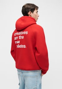 Joven con sudadera roja con texto blanco "los creativos son los nuevos atletas" visto desde atrás, vistiendo jeans azul claro contra un fondo liso.