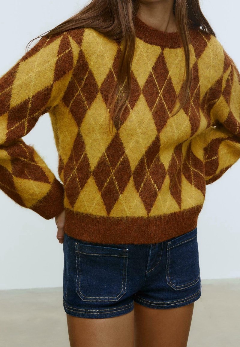 Personne portant un pull en laine jaune et marron à motif losange avec des manches longues et un short en denim bleu foncé.