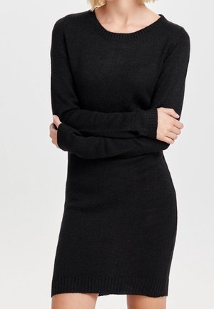 Woman wearing a fitted long-sleeve black knit dress, standing with arms crossed against a plain light background.