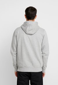 Grijze hoodie met een voorzak, ribbelmanchetten en -zoom. De stof lijkt zacht en textuur te hebben. Gekleed met zwarte broek.