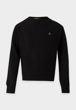 Man draagt een zwart shirt met lange mouwen en klein embleem, losse beige broek en zwarte schoenen, staand tegen een lichte achtergrond.