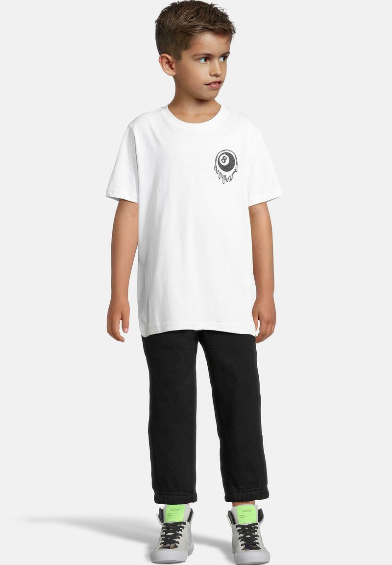 T-shirt en coton blanc à manches courtes, arborant un design d'une boule 8 imprimée en noir sur la poitrine. Associé à un pantalon noir et des baskets gris clair.