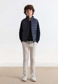 Navy Puffer-Weste über einem schwarzen Pullover, kombiniert mit hellbeigen Pants und weißen Sneakern. Die Weste hat einen hohen Kragen und einen Reißverschluss.