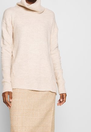 Vaaleanbeige poolopaita, joka on valmistettu pehmeästä neulekankaasta ja jossa on ribbireunat hihansuissa ja helmassa. Yhdistettynä ruudulliseen beigeen hameeseen.