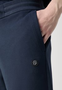 Námornícke bavlnené jogger nohavice s vreckom a embosovaným čiernym logom na gombíku. Hladká textúra so priliehavým elastickým pásom a bočnými švami.