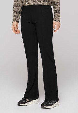 Schwarze, ausgestellte Hosen mit glatter Textur, die ein hoch tailliertes Design aufweisen. Kombiniert mit schwarz-weißen Sportschuhen, geeignet für die Freizeit.