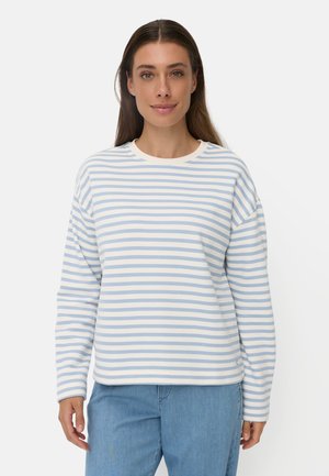 Kvinde med langt brunt hår iført en hvid og lyseblå striped langærmet skjorte og lyseblå jeans, stående mod en ensfarvet baggrund.