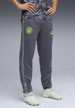 Person iført grå Manchester City træningsbukser med neonlogoer og hvide fodboldstøvler, stående med hænderne på hofterne mod en grå baggrund.