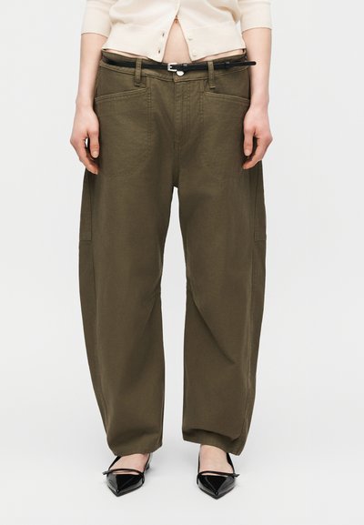 Άτομο που φοράει άνετο χακί πράσινο παντελόνι με μεγάλες μπροστινές τσέπες, λεπτή μαύρη ζώνη, λευκό ζακέτα και μαύρα μυτερά παπούτσια.