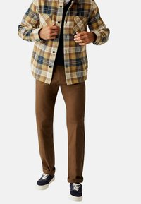 Geruite flanellen blouse in beige, bruin en marineblauw; bruine chino's; marineblauwe sneakers met witte zolen; twee steekzakken aan de voorkant van het shirt.