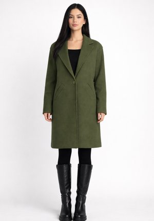 Mujer con cabello largo y oscuro, vestida con un abrigo verde oliva hasta la rodilla, leggings negros y botas negras hasta la rodilla, de pie contra un fondo blanco.