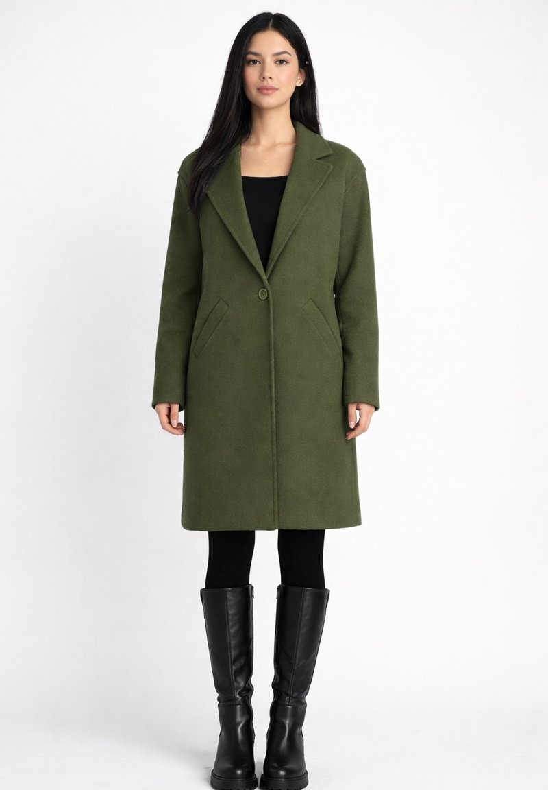 Vrouw met lang donker haar die een olijfgroene jas op knielengte draagt, zwarte legging en zwarte laarzen tot de knie, staande tegen een witte achtergrond.