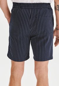 Granatowe szorty z białymi pionowymi paskami, z elastycznym paskiem w talii i tylną kieszenią. Materiał wydaje się gładki i lekki.