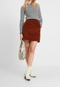 Mini gonna incordata marrone, top a maniche lunghe bianco e nero a righe, borsa a tracolla bianca con stampa serpente, e stivali alla caviglia crema con tacco a blocco.