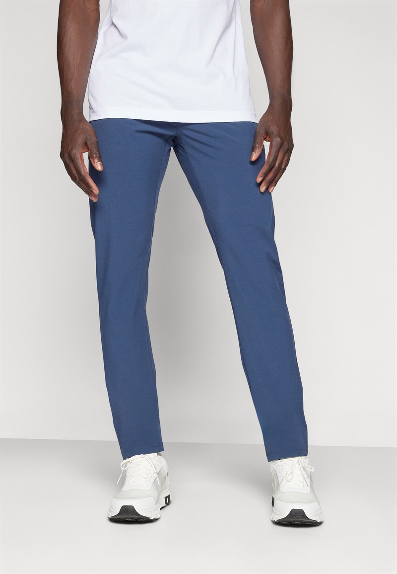 Blaue, slim-fit Hosen aus dehnbarem Stoff mit glatter Textur und ohne sichtbare Muster oder Beschläge. Kombiniert mit weißen Sneakers.