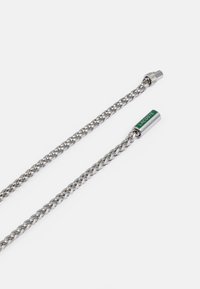 Corrente de metal prateado com design trançado, apresentando um fecho cilíndrico com o logótipo verde "LACOSTE". Textura suave e acabamento brilhante.