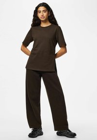 Femme aux longs cheveux bouclés portant un t-shirt ample marron foncé et un pantalon, avec des chaussures de course noires, debout les mains derrière le dos.