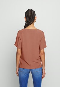 Blouse à manches courtes couleur rouille avec encolure bateau, coupe décontractée et revers de manches roulés, associée à un jean en denim bleu.