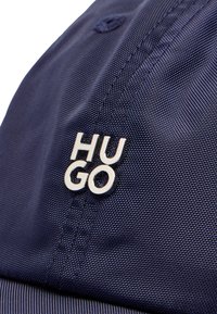 Gorra azul marino hecha de tela texturizada, con un logotipo plateado pulido "HUGO" en la parte delantera. Incluye un detalle de costura.