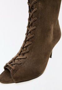 Bottine marron en daim à talon haut avec un laçage devant et un bout ouvert sur fond blanc.