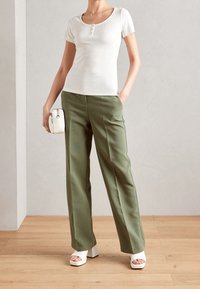 Valkoinen lyhythihainen paita nappidetaljeilla, yhdistetty oliivinvihreisiin leveälahkeisiin housuihin. Malli pitää kädessään pientä valkoista käsilaukkua ja käyttää valkoisia korollisia sandaaleja.